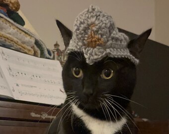 May include: A black cat with white markings and a gray crocheted hat with a brown accent sits on a wooden surface. The cat is in front of a piano with sheet music. The cat has white paws and a white patch on its chest.
