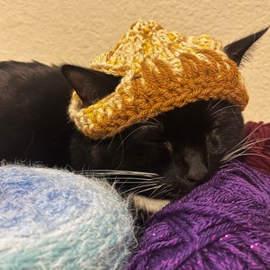 May include: A black cat wearing a crocheted brown and yellow hat, sleeping on a pile of colourful yarn.