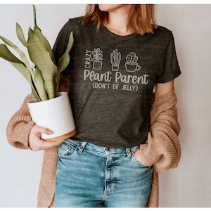 May include: A dark gray t-shirt with the text "Crazy Plant Parent (Don't Be Jelly)" and a graphic of three potted plants. The person wearing the shirt is holding a potted plant.