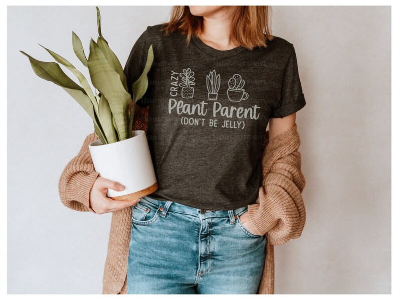 May include: A dark gray t-shirt with the text "Crazy Plant Parent (Don't Be Jelly)" and a graphic of three potted plants. The person wearing the shirt is holding a potted plant.