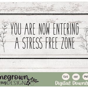 Pode incluir: Uma placa preta e branca com o texto "YOU ARE NOW ENTERING A STRESS FREE ZONE" com desenhos florais de cada lado. A placa está sobre um fundo de madeira branca.