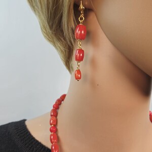 May include: A pair of drop earrings featuring three graduated, smooth, cylindrical red coral beads linked by gold-toned metal, paired with a matching necklace of similar red coral beads worn against a black top.