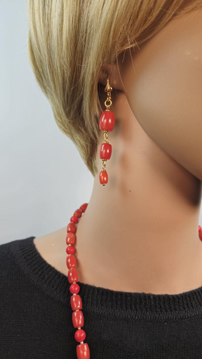 May include: A pair of drop earrings featuring three graduated, smooth, cylindrical red coral beads linked by gold-toned metal, paired with a matching necklace of similar red coral beads worn against a black top.