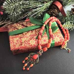 May include: A necklace featuring smooth, cylindrical coral-colored beads interspersed with small gold-toned spacers and a central ornate pendant. The pendant has a teardrop-shaped coral stone, intricate black and gold metalwork, and dangling bead accents.