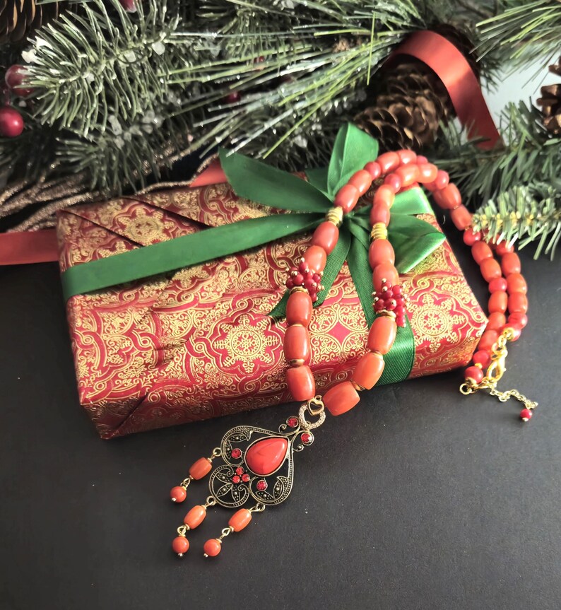 May include: A necklace featuring smooth, cylindrical coral-colored beads interspersed with small gold-toned spacers and a central ornate pendant. The pendant has a teardrop-shaped coral stone, intricate black and gold metalwork, and dangling bead accents.