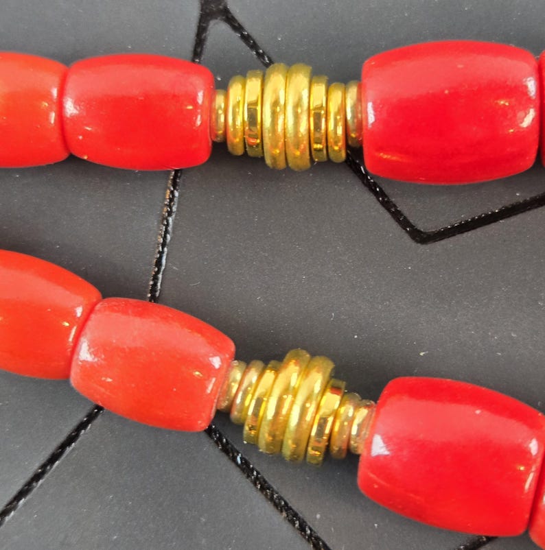 May include: A necklace featuring smooth, cylindrical red beads alternating with smaller, ribbed gold-tone metal spacers. The beads are arranged in a repeating pattern, showcasing a vibrant red color contrasted by the metallic gold accents throughout.