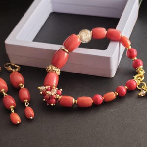May include: A jewelry set consisting of a bracelet and two drop earrings. The bracelet features cylindrical coral-colored beads, gold-tone spacers, a clear bead, and a cluster of small red and clear beads. The earrings have matching coral beads.