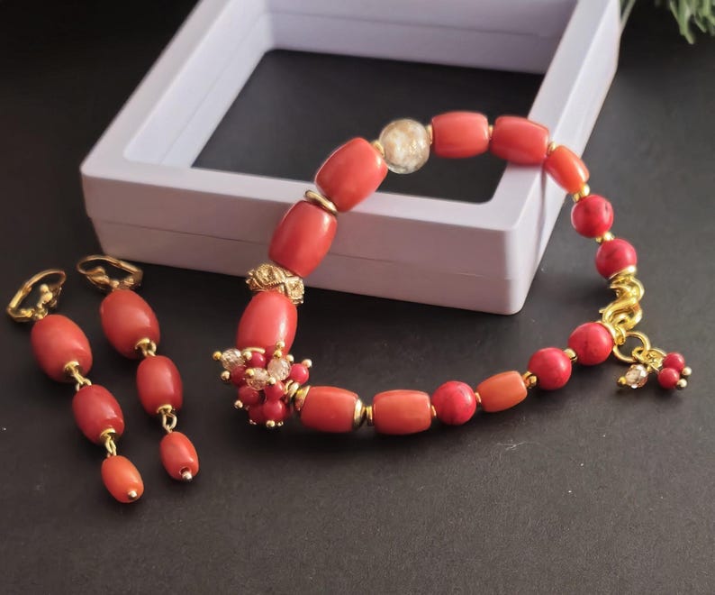 May include: A jewelry set consisting of a bracelet and two drop earrings. The bracelet features cylindrical coral-colored beads, gold-tone spacers, a clear bead, and a cluster of small red and clear beads. The earrings have matching coral beads.