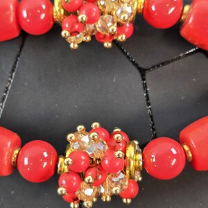 May include: A bracelet featuring smooth, bright red cylindrical and spherical beads interspersed with clusters of small red beads, clear faceted crystals, and gold-toned metal accents. The design includes repeating segments of these vibrant red components.
