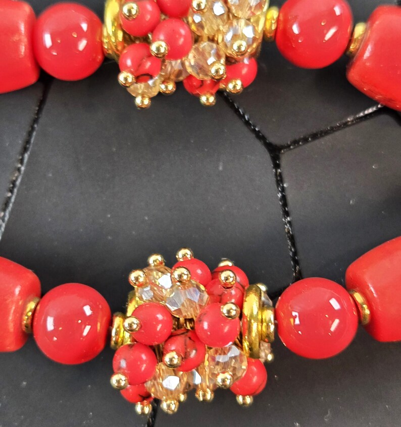 May include: A bracelet featuring smooth, bright red cylindrical and spherical beads interspersed with clusters of small red beads, clear faceted crystals, and gold-toned metal accents. The design includes repeating segments of these vibrant red components.