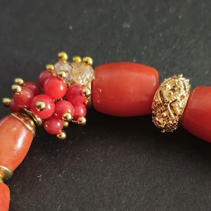 May include: A bracelet featuring smooth, cylindrical red beads interspersed with clusters of smaller, round red beads and gold-toned metal accents. The design includes a textured, filigree-style gold bead and small, clear faceted crystal beads.