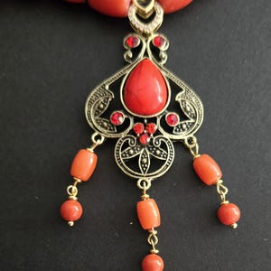 May include: A necklace featuring a series of orange cylindrical beads and a central ornate gold-tone metal pendant. The pendant includes a large red teardrop-shaped stone, smaller red accent stones, and three dangling strands of orange beads.