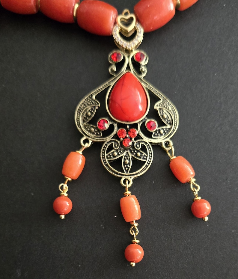 May include: A necklace featuring a series of orange cylindrical beads and a central ornate gold-tone metal pendant. The pendant includes a large red teardrop-shaped stone, smaller red accent stones, and three dangling strands of orange beads.