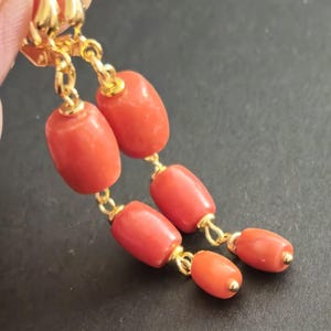 May include: A pair of drop earrings featuring polished, cylindrical, coral-colored beads suspended from gold-toned metal hooks. Each earring consists of two vertically aligned beads connected by small gold-toned jump rings, creating a dangling design.