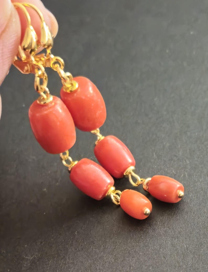 May include: A pair of drop earrings featuring polished, cylindrical, coral-colored beads suspended from gold-toned metal hooks. Each earring consists of two vertically aligned beads connected by small gold-toned jump rings, creating a dangling design.