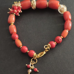 May include: A bracelet featuring a mix of cylindrical and spherical red coral beads, interspersed with gold-toned metal spacers, a textured gold-toned bead, and a clear glass bead with gold flecks, finished with a gold-toned lobster clasp.