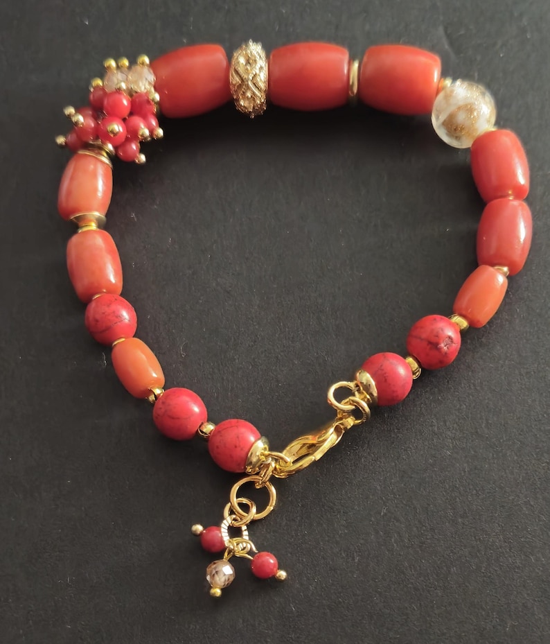 May include: A bracelet featuring a mix of cylindrical and spherical red coral beads, interspersed with gold-toned metal spacers, a textured gold-toned bead, and a clear glass bead with gold flecks, finished with a gold-toned lobster clasp.