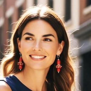 May include: A pair of drop earrings featuring smooth, cylindrical red beads accented with smaller, textured red floral-shaped components. The earrings are suspended from gold-toned metal hooks and hang vertically from the earlobes.