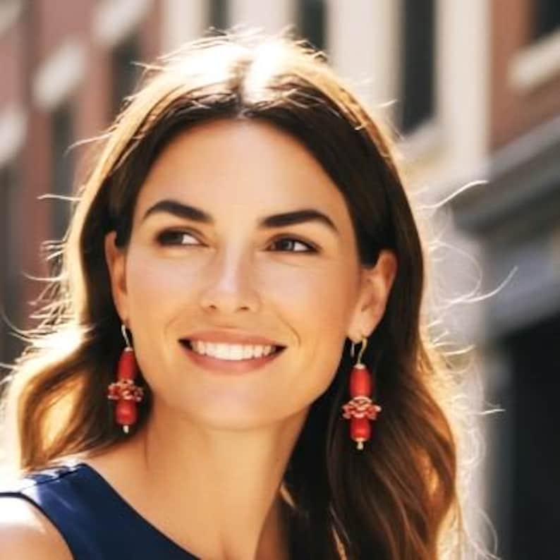 May include: A pair of drop earrings featuring smooth, cylindrical red beads accented with smaller, textured red floral-shaped components. The earrings are suspended from gold-toned metal hooks and hang vertically from the earlobes.