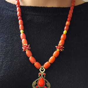 May include: A necklace featuring smooth, cylindrical orange-red beads interspersed with small gold-toned spacers and clusters of tiny red beads. A central gold-toned filigree pendant holds a teardrop-shaped orange-red stone and small red accents.