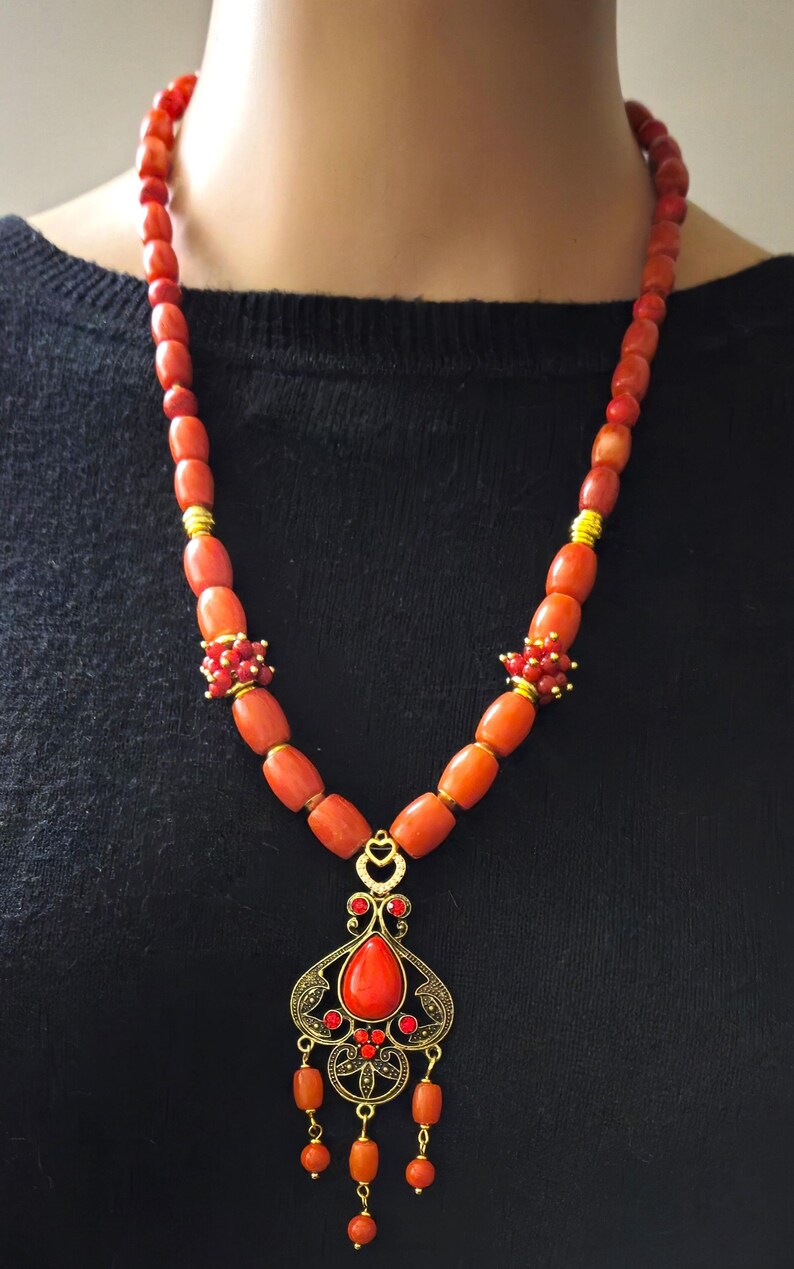 May include: A necklace featuring smooth, cylindrical orange-red beads interspersed with small gold-toned spacers and clusters of tiny red beads. A central gold-toned filigree pendant holds a teardrop-shaped orange-red stone and small red accents.