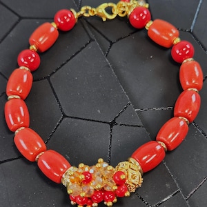 May include: A bracelet featuring smooth, cylindrical red beads interspersed with small gold-toned spacers. The piece includes a central cluster of tiny red beads and clear crystals, finished with a gold-toned lobster claw clasp closure.