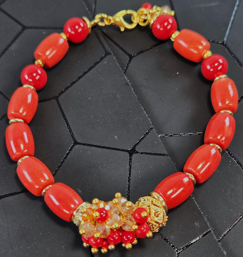 May include: A bracelet featuring smooth, cylindrical red beads interspersed with small gold-toned spacers. The piece includes a central cluster of tiny red beads and clear crystals, finished with a gold-toned lobster claw clasp closure.