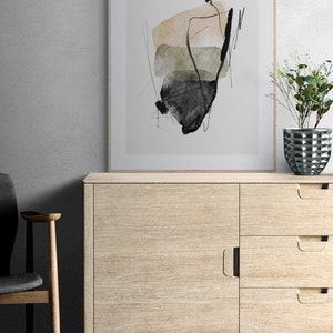 May include: A framed abstract art print with black, brown, and green watercolor shapes on a white background. The print is hanging on a white wall above a light wood cabinet with drawers and doors.
