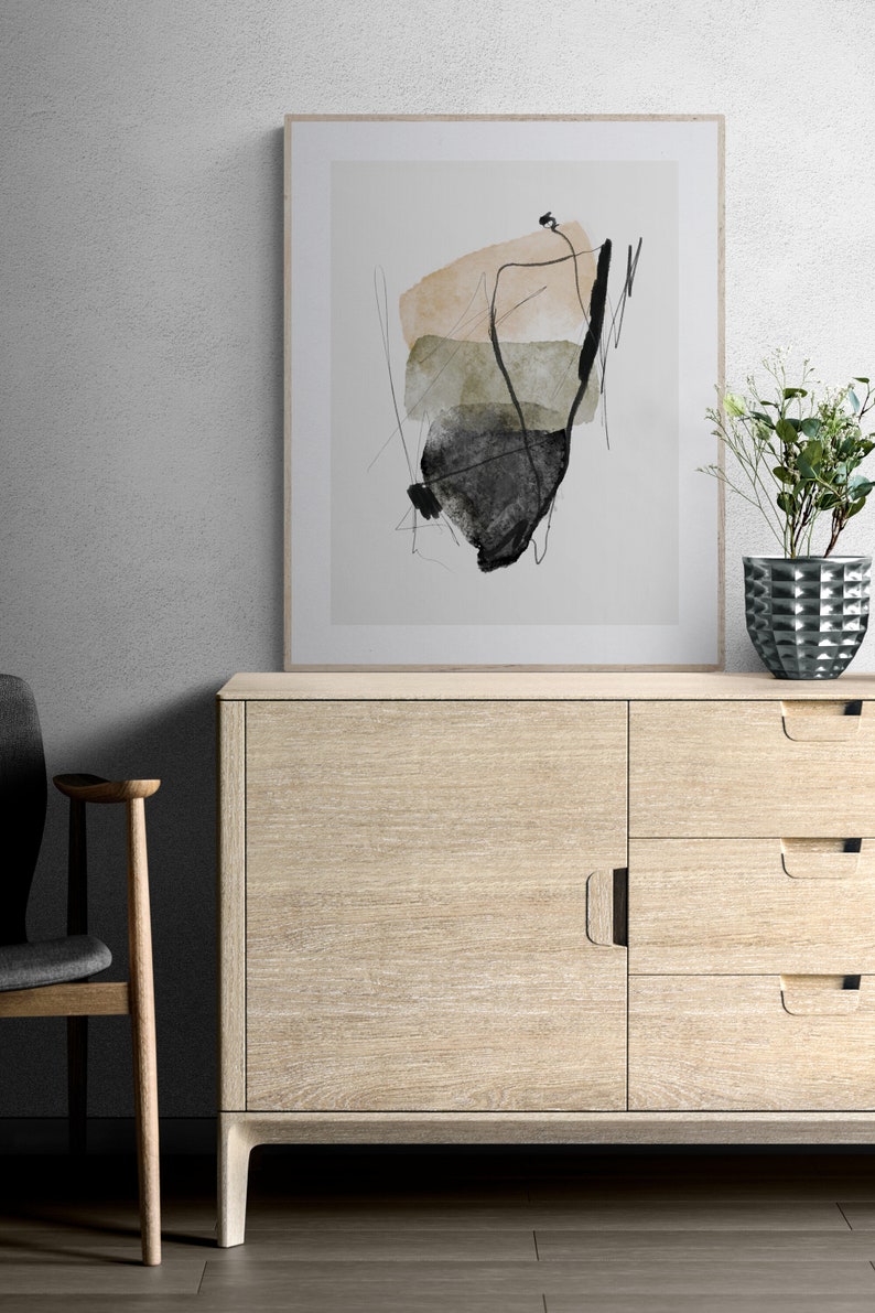 May include: A framed abstract art print with black, brown, and green watercolor shapes on a white background. The print is hanging on a white wall above a light wood cabinet with drawers and doors.