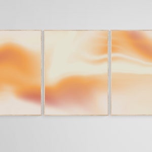 May include: Three framed abstract art prints with a soft, blurred, orange and white colour scheme. The prints are arranged in a horizontal row.