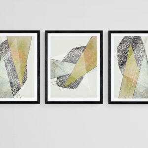 Op de afbeelding: Een set van drie abstracte kunstprints in zwarte frames. Elke print heeft een geometrisch ontwerp met overlappende vormen in tinten groen, grijs en wit.