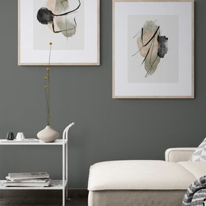 May include: Two framed abstract art prints with black lines and green and beige watercolor shapes on a white background. The prints are hanging on a dark green wall above a white couch.