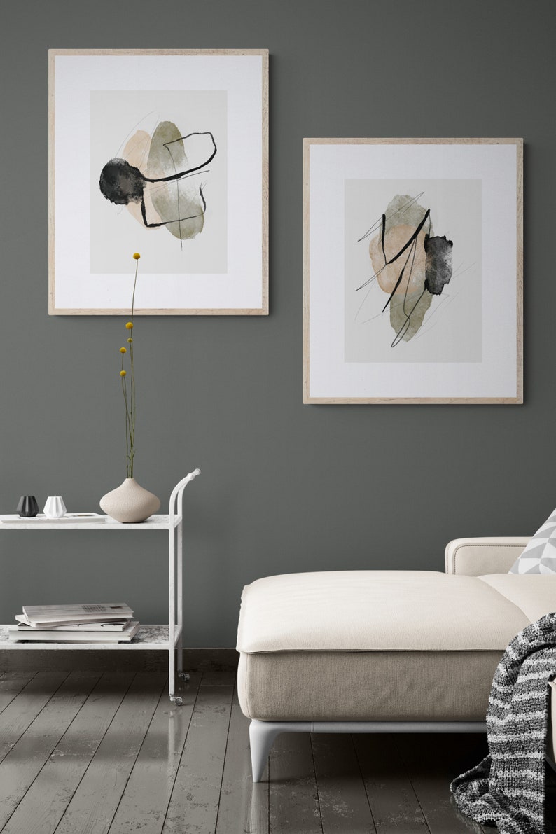 May include: Two framed abstract art prints with black lines and green and beige watercolor shapes on a white background. The prints are hanging on a dark green wall above a white couch.
