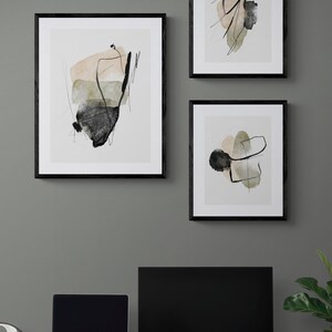 May include: Three black framed abstract art prints with black and white lines and watercolor washes in shades of beige, green, and gray. The prints are hanging on a gray wall above a desk with a laptop, monitor, and other office supplies.