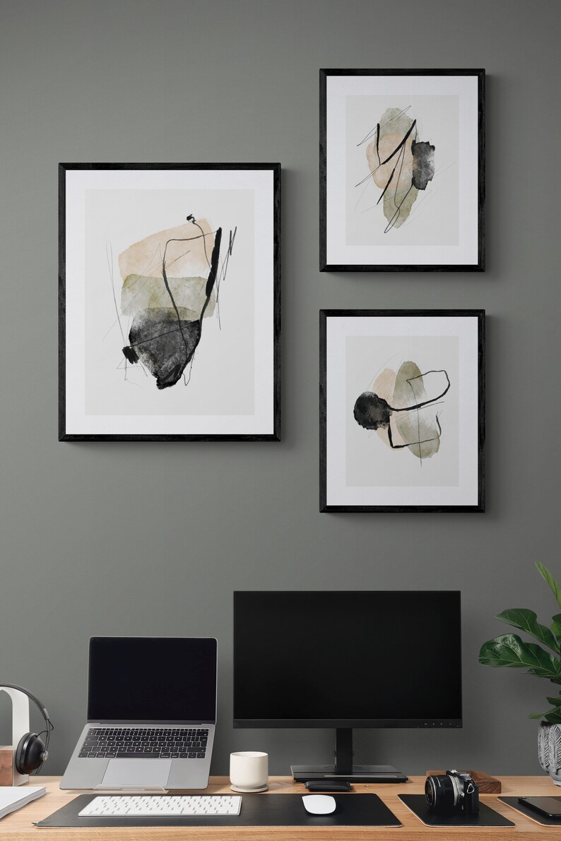 May include: Three black framed abstract art prints with black and white lines and watercolor washes in shades of beige, green, and gray. The prints are hanging on a gray wall above a desk with a laptop, monitor, and other office supplies.