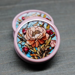 May include: A pair of pink ear gauges with a stained glass floral design featuring a large pink flower and smaller red and pink flowers. The design is set against a blue and white background.