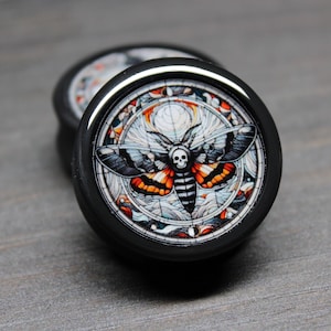 May include: A pair of black ear gauges with a circular design featuring a detailed illustration of a moth with a skull on its body. The moth is surrounded by a circular pattern of geometric shapes and lines.