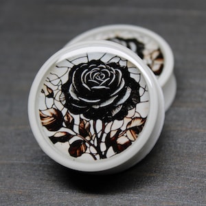 May include: White ear gauges with a black rose design. The rose is detailed with dark brown leaves and a white background. The gauges are round and have a smooth, glossy finish.