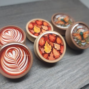 May include: Six round wooden coasters with different designs.  Two coasters feature a latte art design, two coasters feature a fall leaf design, and two coasters feature a butterfly design.
