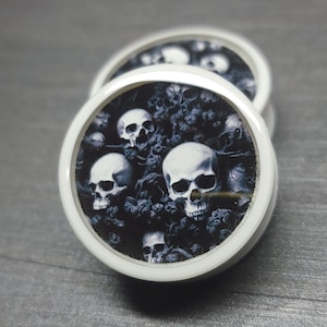 May include: Pair of white ear gauges with a black and white skull design. The design features multiple skulls with a dark background.