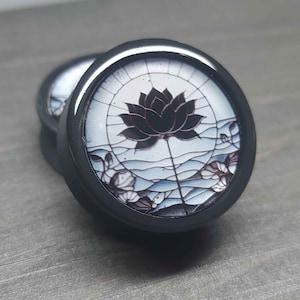 May include: Black ear gauges with a detailed lotus flower design. The image features two gauges, each with a black frame and a central image of a dark lotus against a light blue and white background, evoking a stained-glass effect.