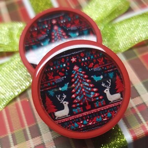 May include: Two red round Christmas ornaments with a white and blue patterned design featuring a Christmas tree and reindeer. The ornaments are on a red and green plaid background.