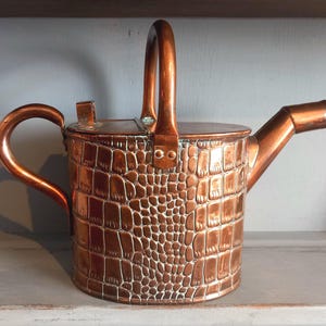 May include: A polished copper watering can featuring a textured crocodile skin pattern on the cylindrical body. It includes a curved side handle, a vertical top handle, a flat lid with a small tab, and a short, angled spout extending to the right.