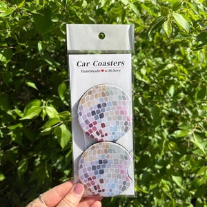 May include: Two round car coasters with a mosaic tile design in shades of gray, blue, pink, and brown. The coasters are packaged in a clear plastic bag with a white label that reads "Car Coasters Handmade with love".