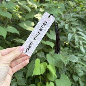 May include: A rectangular bookmark with the text "DEAREST GENTLE READER" in black. The bookmark has a white lace pattern on a pink background and a black tassel. The background is a lush green foliage.