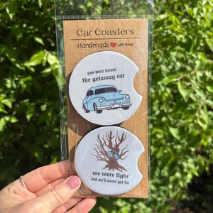May include: Two round car coasters with a white background and black text. The top coaster features a blue car with the text "you were drivin' the getaway car". The bottom coaster features a brown tree with blue eyes and the text "we were flyin' but we'd never get far".