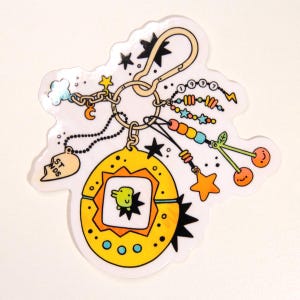 May include: A yellow and orange charm with a black star and a small green creature inside a square. The charm is attached to a silver chain with a heart charm that says "ST. NDS" and a silver ring with a crescent moon charm. The charm is also attached to a gold key ring with a variety of colorful beads, stars, and cherries.