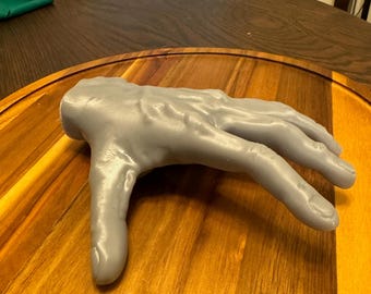 May include: A grey 3D printed hand model with realistic detail. The hand is posed in a relaxed, open position with fingers slightly spread apart. The model is resting on a wooden surface.