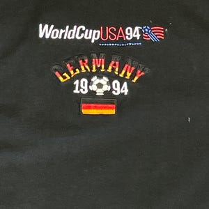 May include: Black t-shirt with the text "World Cup USA 94" and a small American flag. Below the text is a curved graphic with the text "Germany 19 94" and a soccer ball. A small German flag is below the text.