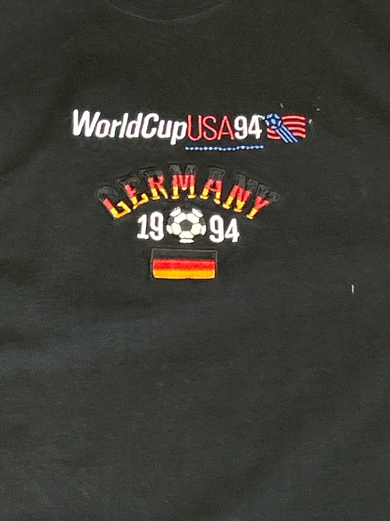 May include: Black t-shirt with the text "World Cup USA 94" and a small American flag. Below the text is a curved graphic with the text "Germany 19 94" and a soccer ball. A small German flag is below the text.